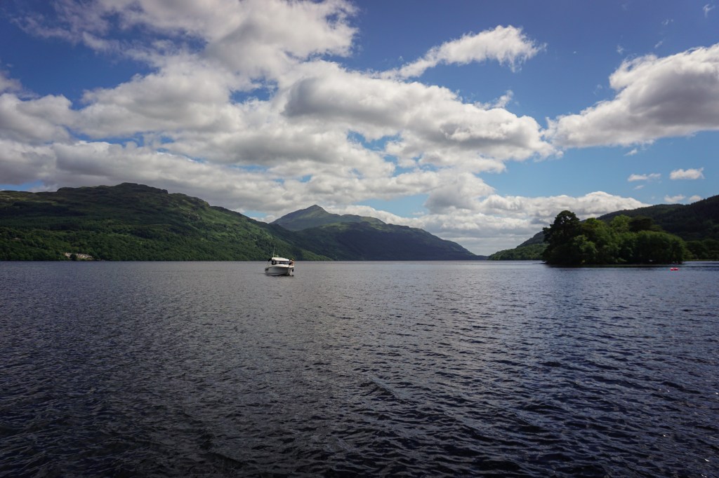 Looking over Loch Lomond, there are some mountains in the distance and a small white boat coming towards the camera