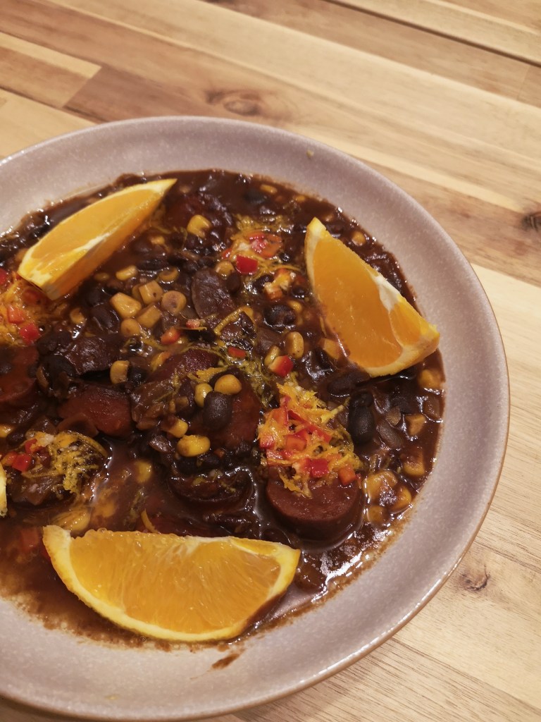 A bowl of a brown looking mince and chorizo stew called feijoada, there are wedges of orange on top