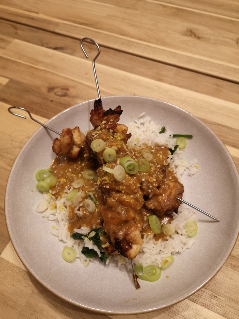 Two chicken skewers covered in satay sauce over a bed of white rice and topped with spring onions