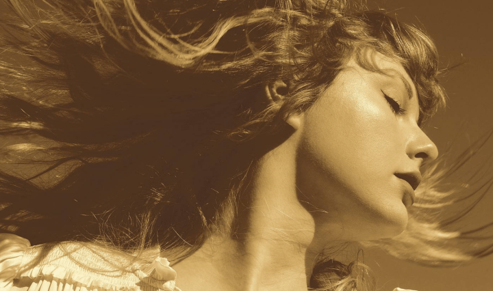 Taylor Swift looks to the right, her hair splaying out from the movement, her eyes are closed and she is wearing a frilly white blouse. The photo has a sepia style filter on it