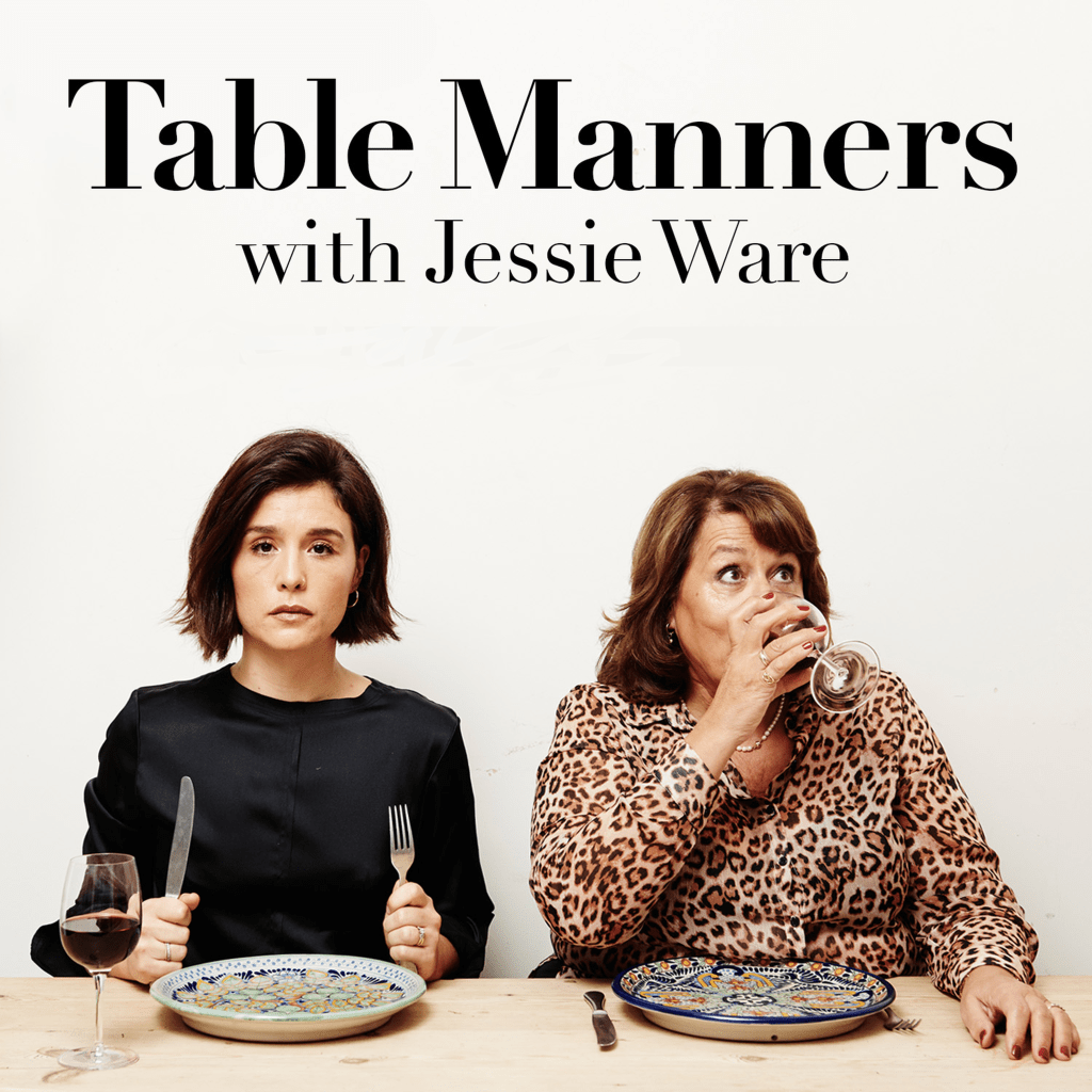 Two women sit at a table, the younger holds up her fork and knife with a straight face, the older woman drinks her glass of red wine with wide eyes looking up