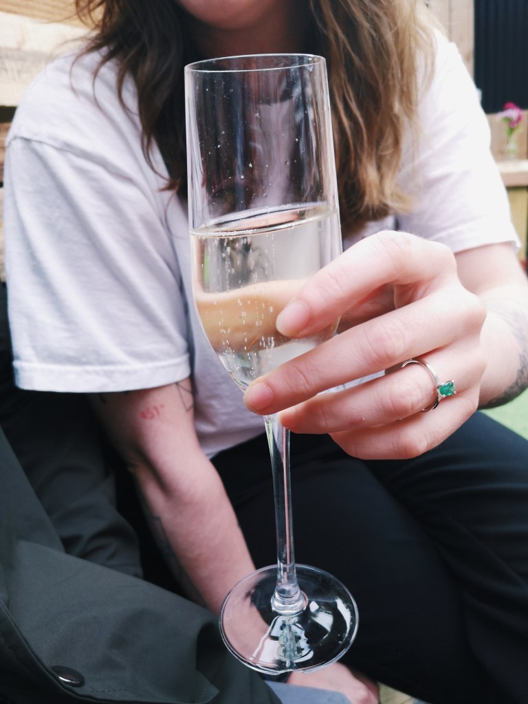 A woman's hand holds a flute glass of fizzing prosecco with a silver emerald ring on her finger