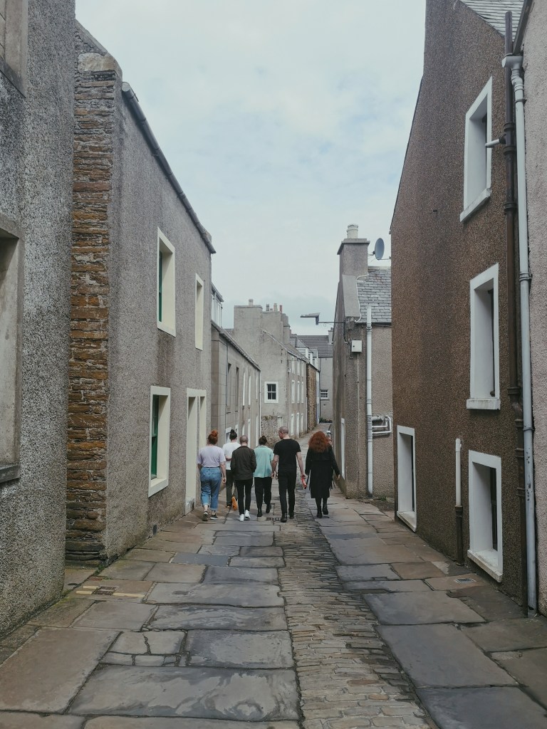 Six people are walking down a wee lane surrounded by little grey houses