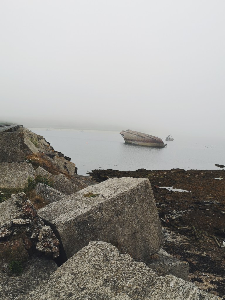 A pile of square grey stones are lined up along the side and in the distance we can see a boat lying on it's side halfway in the water