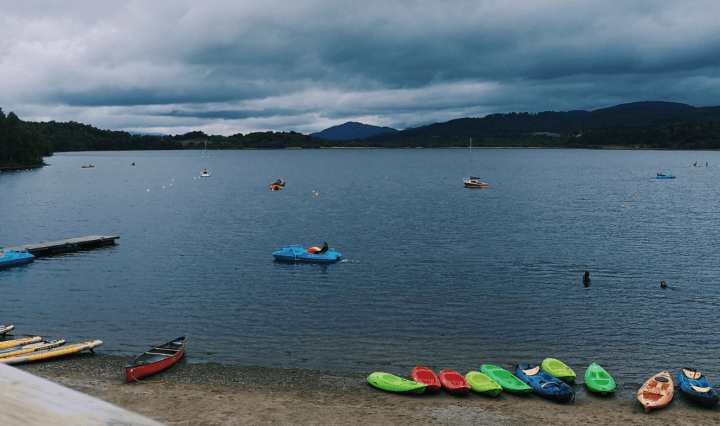 A large loch stretches to the distance, there are various kayaks, boats and paddle boards on the shore and in the water