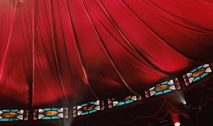 The roof of a circus tent, red material drapes down toward a row of stain glass window panels
