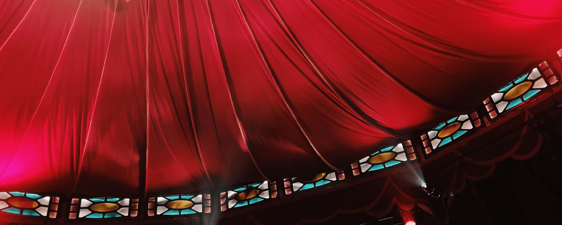 The roof of a circus tent, red material drapes down toward a row of stain glass window panels