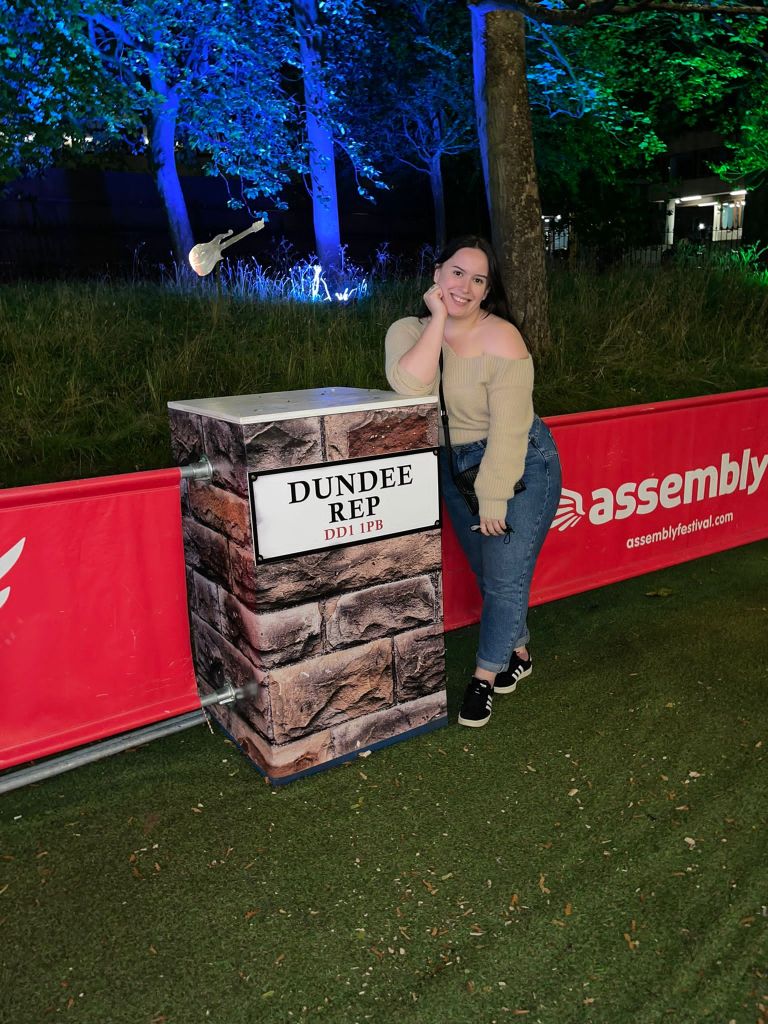 A young woman stands leaning on a table that has a sign saying Dundee Rep