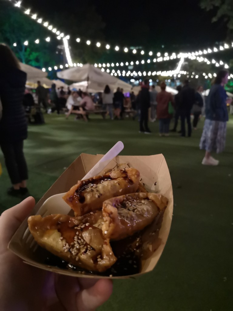 Three gyoza dumplings in a little cardboard tray, in the background people sit at tables under umbrellas and there are fairy lights strung up above
