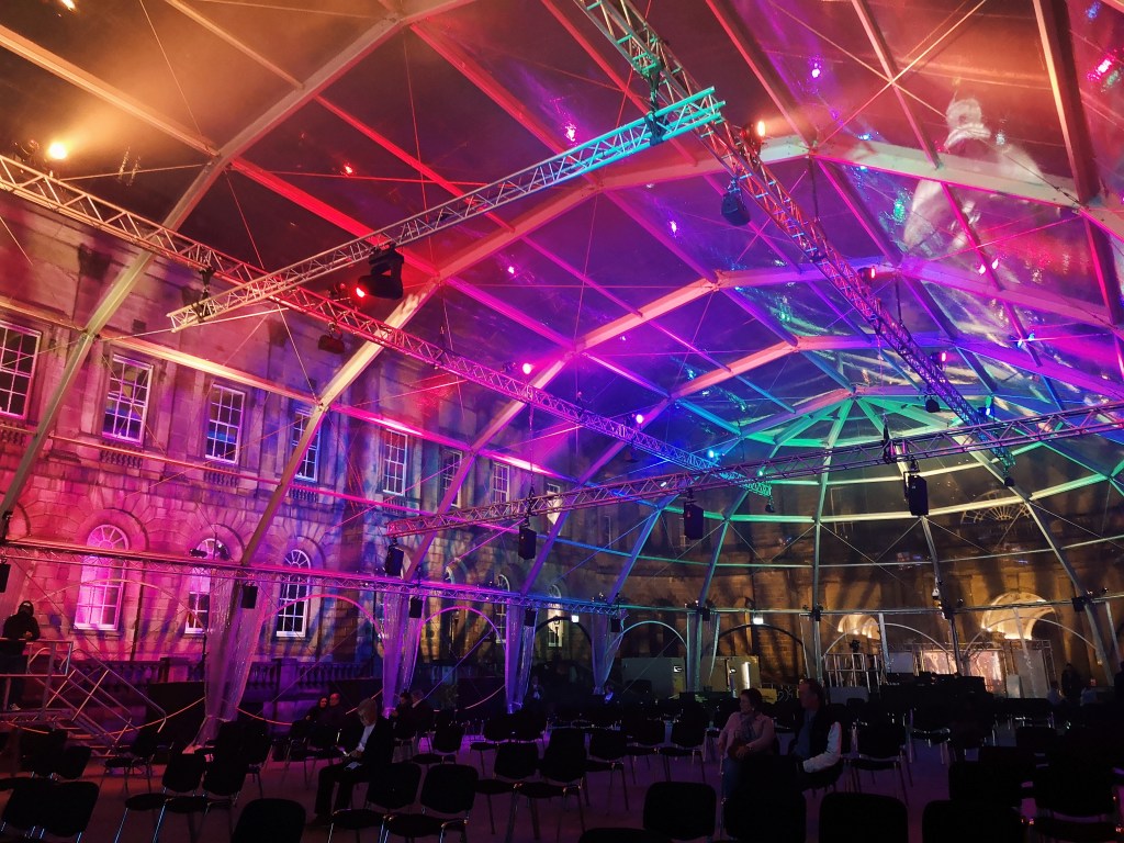 A large arched structure in a courtyard of old buildings, the big tent is transparent and glowing with various coloured lights