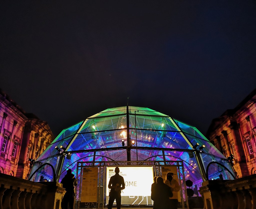 A large transparent dome placed between two old buildings, everything is lit with colourful lights