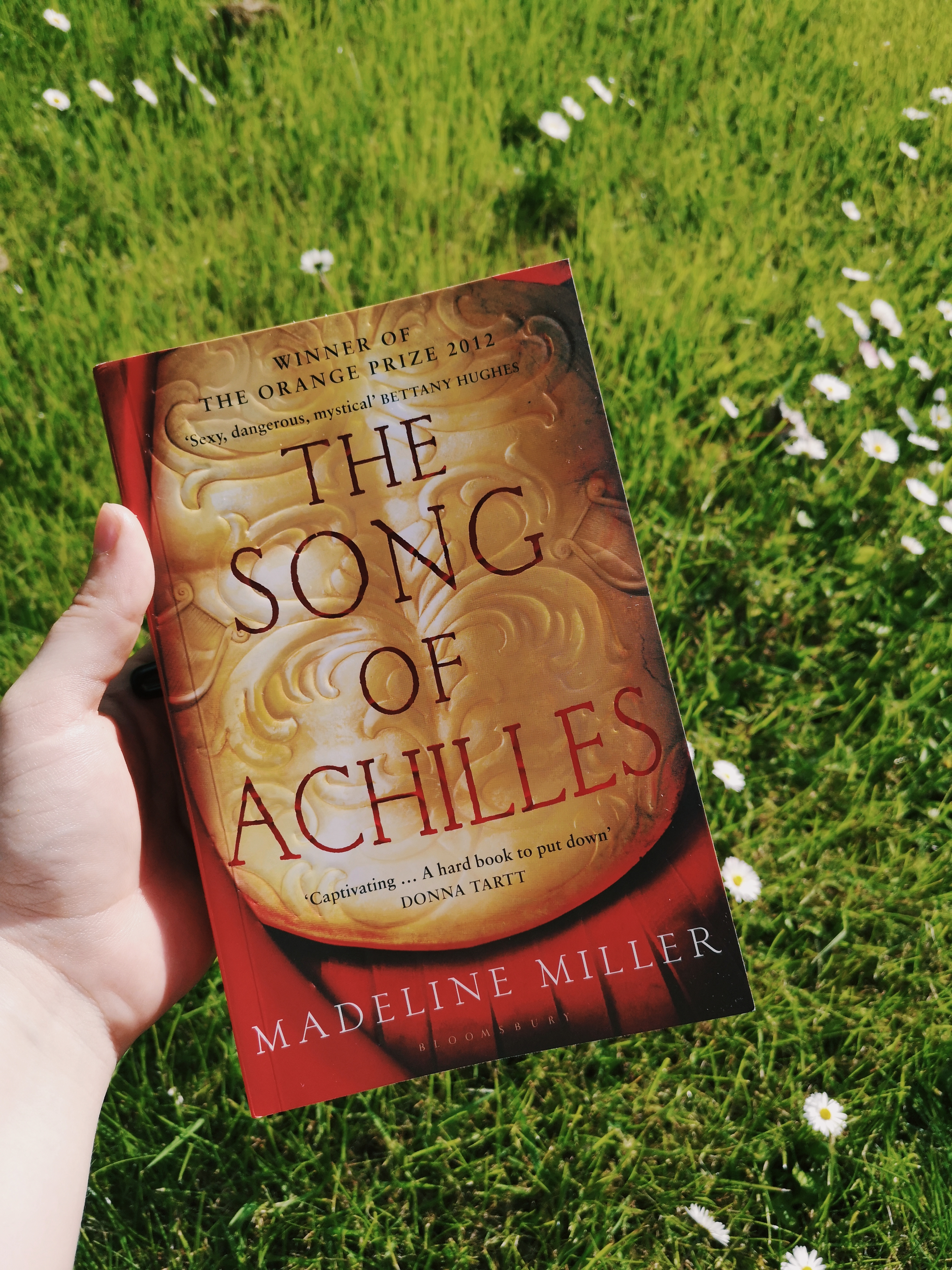 A hand holds the book The Song of Achilles by Madeline Miller in front of green grass with daisies