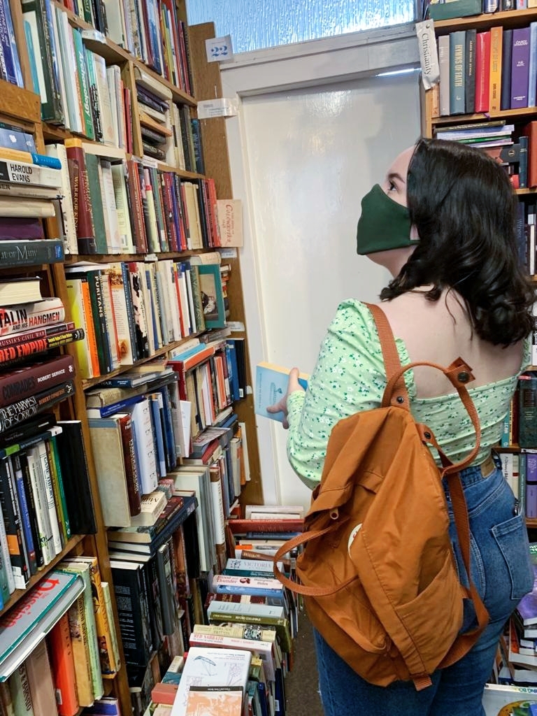 A woman looks up at a wall of bookshelves packed with books, she is wearing a face mask, jeans, a green floral top and is holding an orange bagpack and a book in her hands