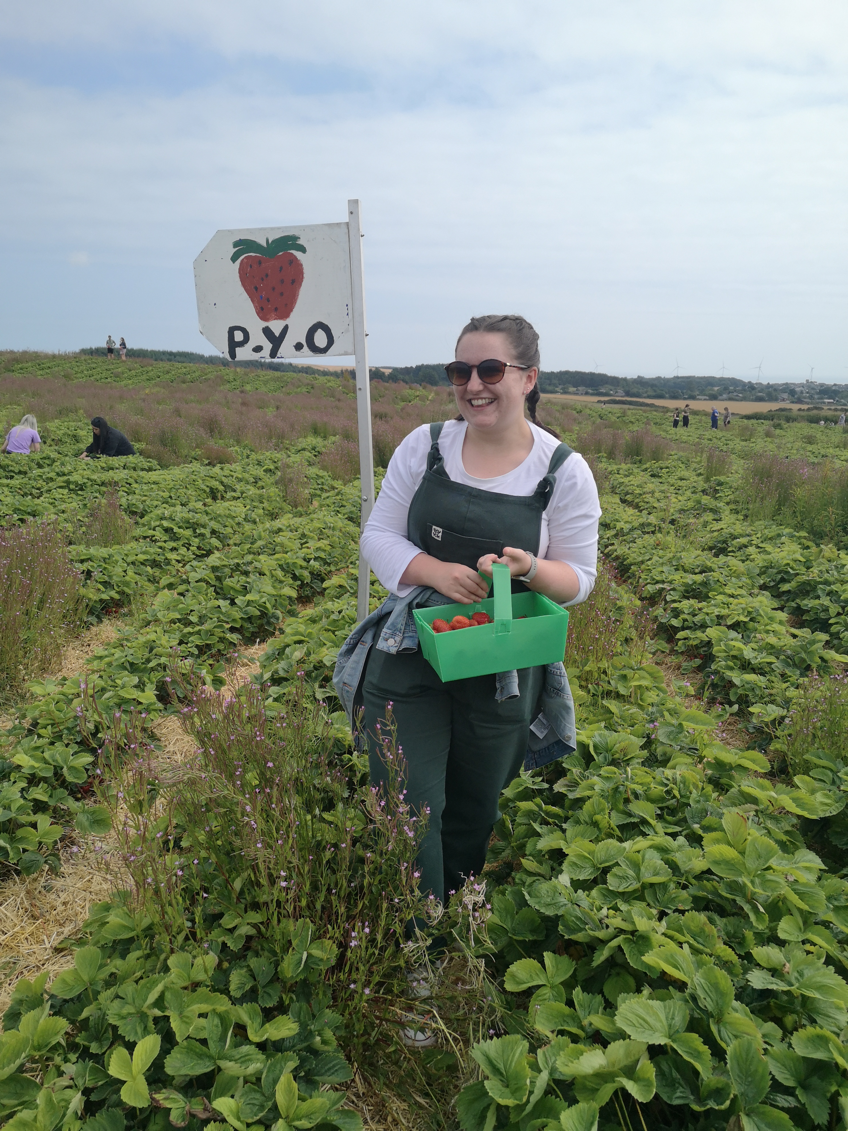 A young woman in green dungarees stands in a field holding a punnet of strawberries, behind her is a sign that says PYO (Pick Your Own)