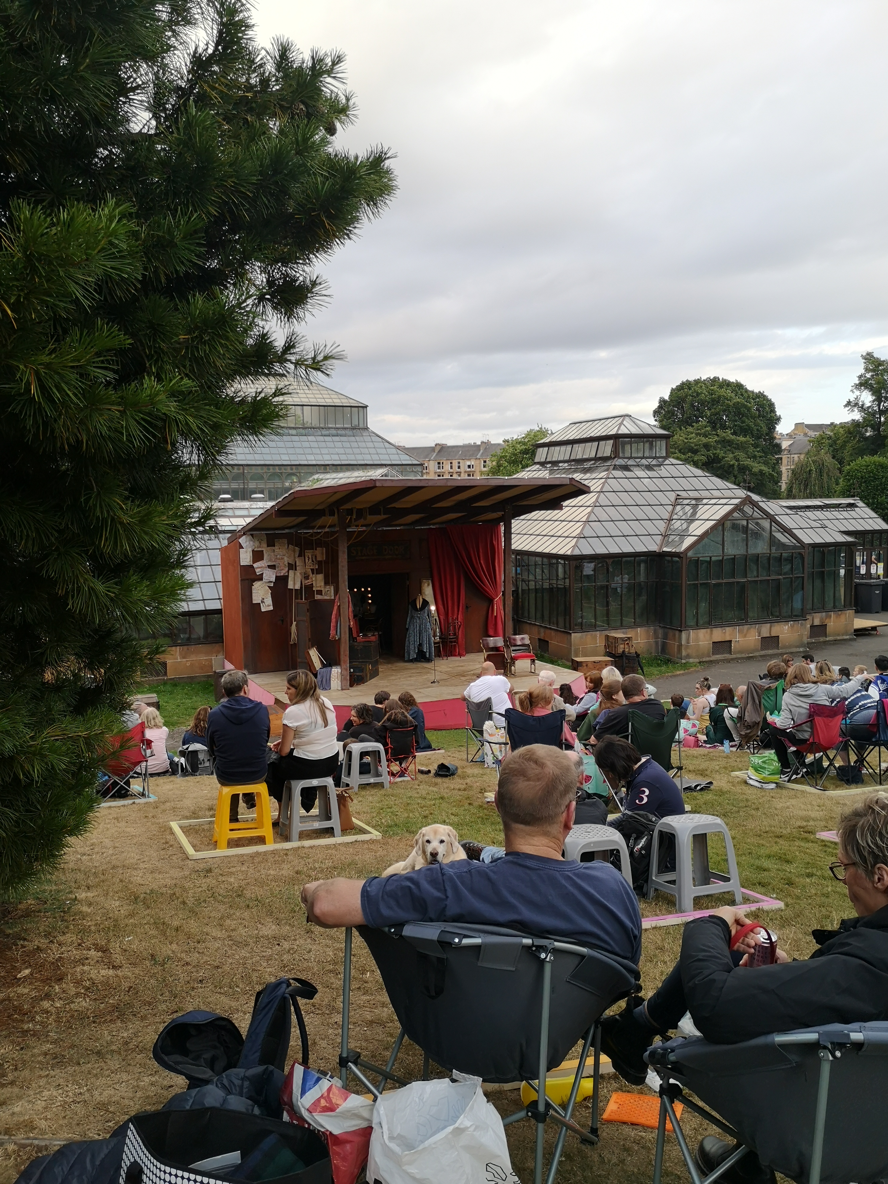 A small stage is set up in a park with people sitting on the grass spaced out