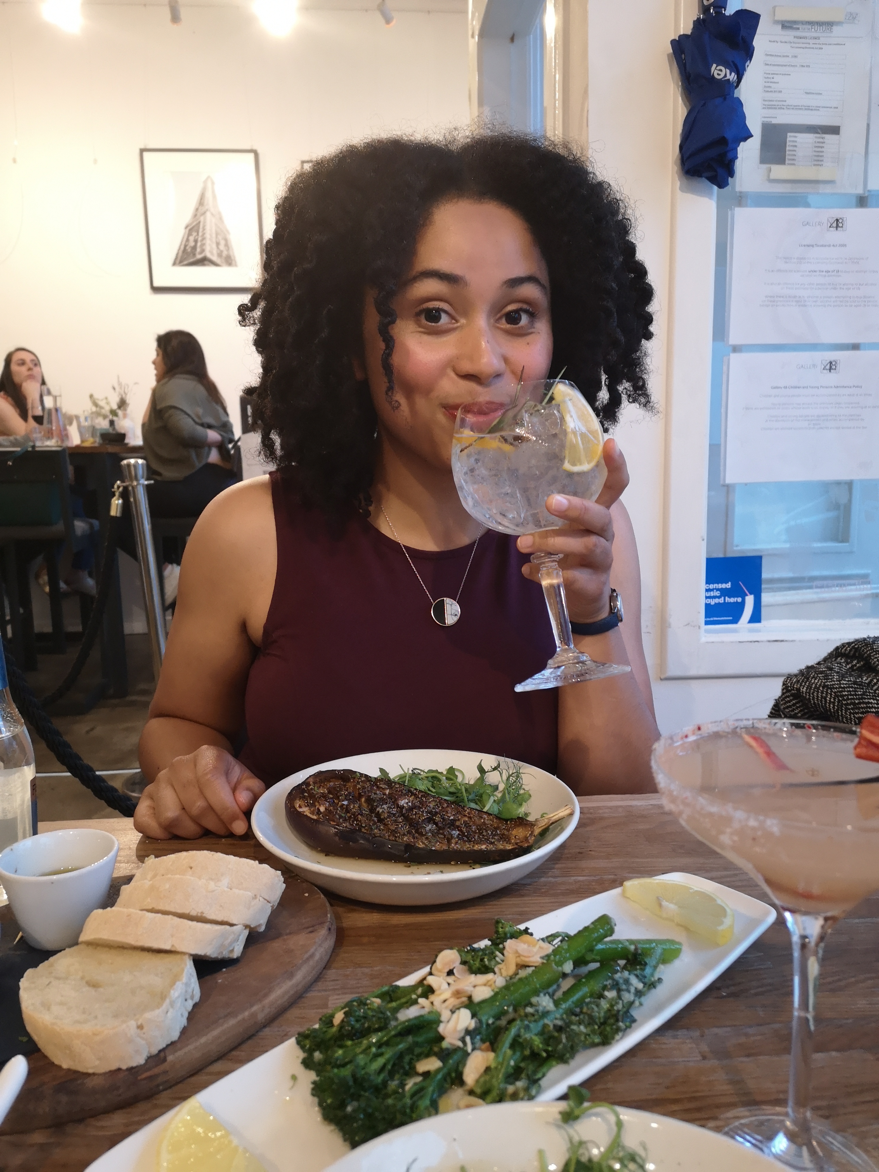 A woman holds a gin and tonic to her lips and smiles, there is food on the table in front of her