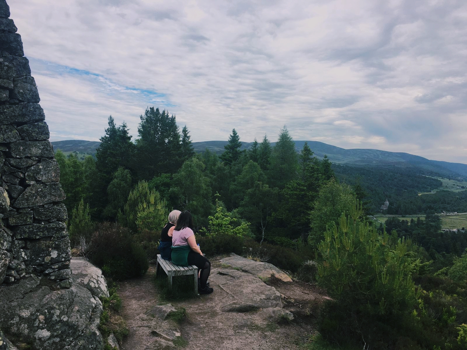 Me and my mum sit on a bench on top of a hill overlooking Scottish countryside, covered with trees