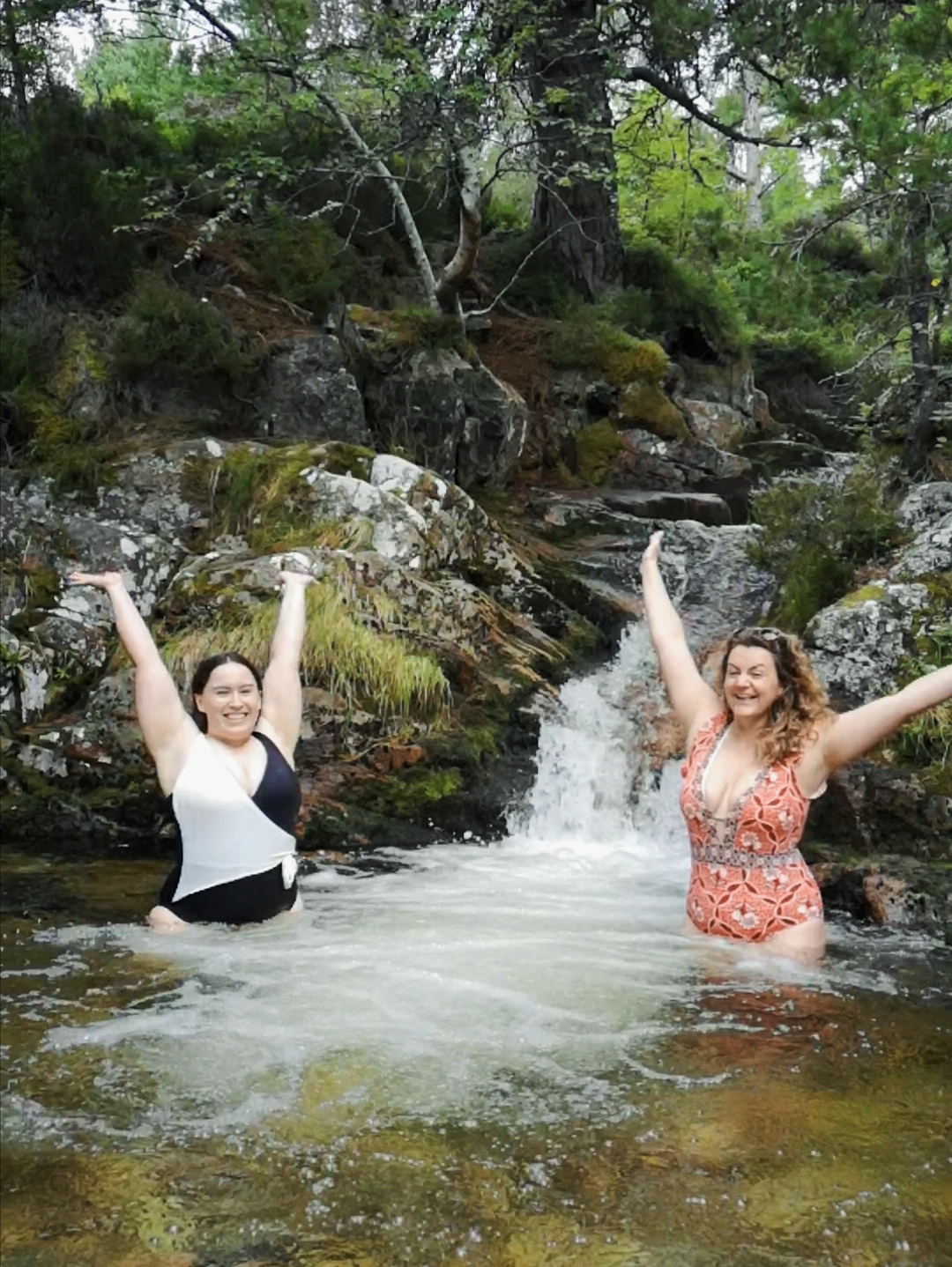 Two women stand in a brook beside a waterfall, they have their arms stretched up overhead