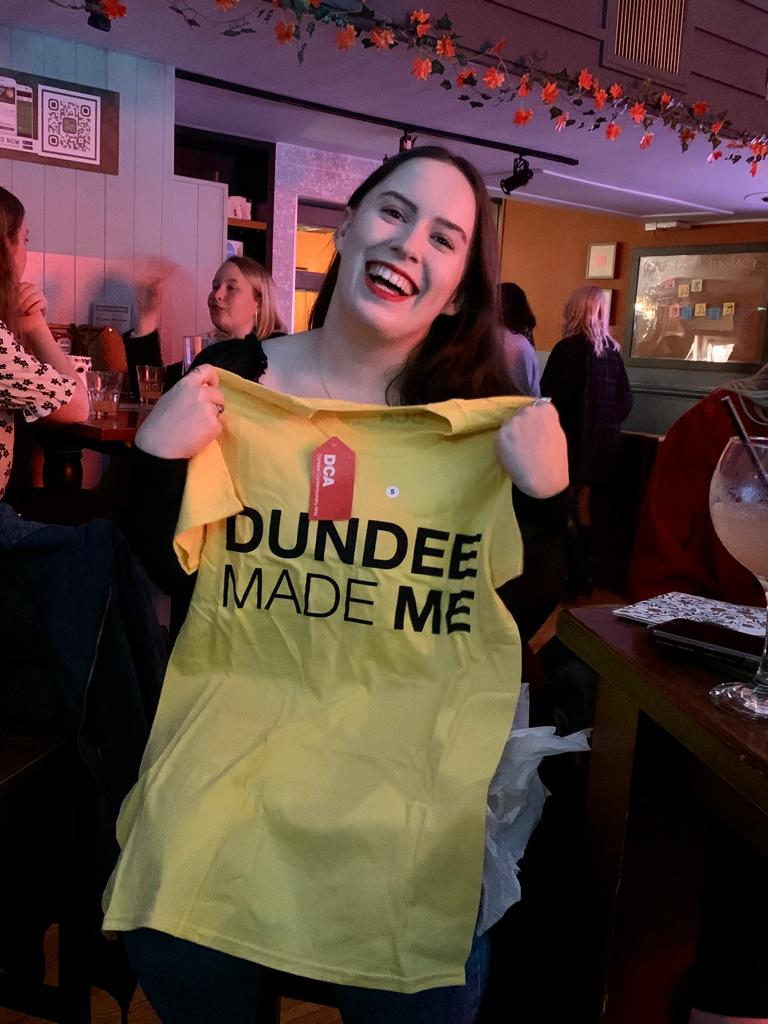 A woman smiles while holding up a tshirt which says 'Dundee Made Me'