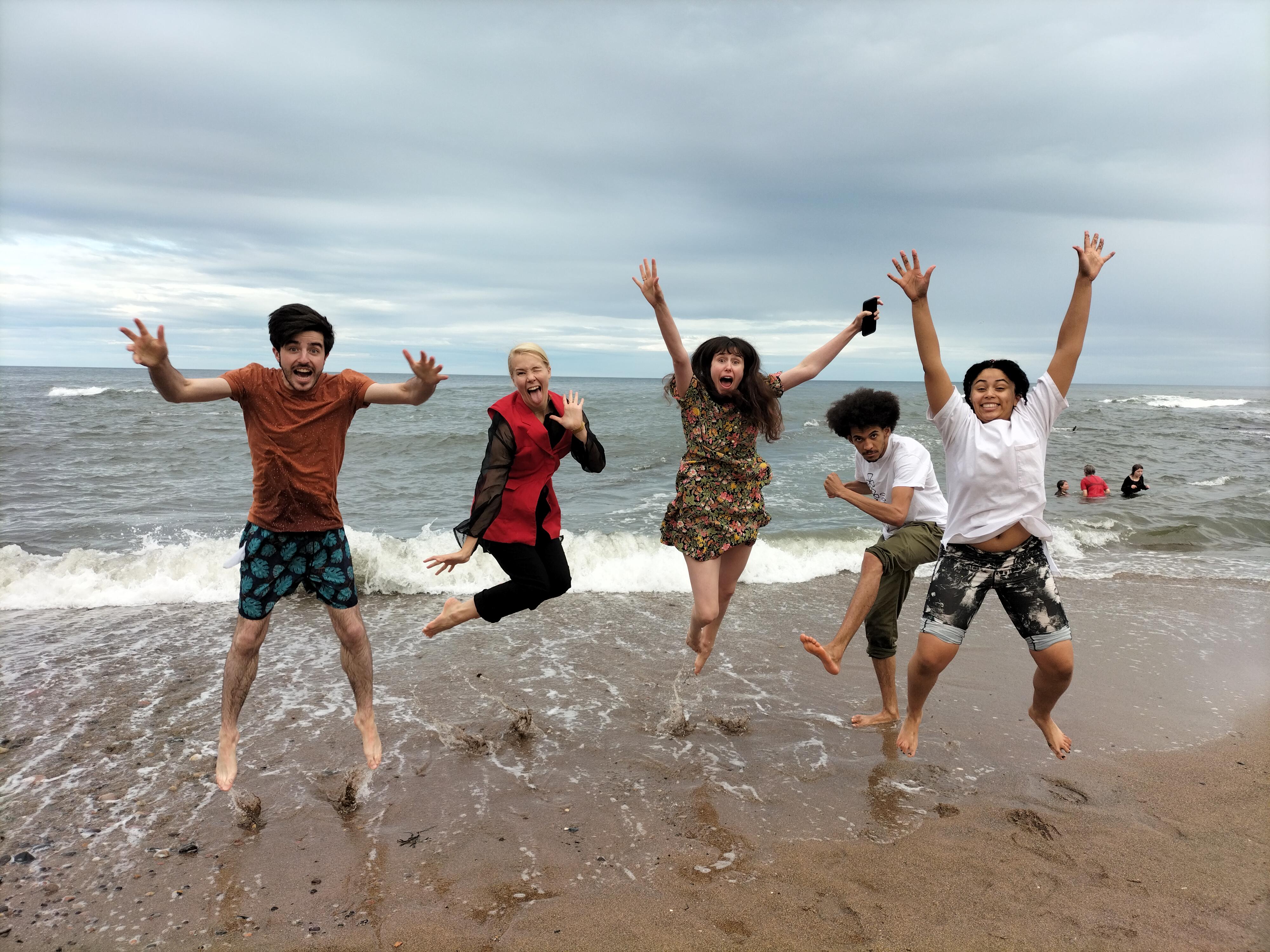 Five people on a beach try to jump at the same time and they make crazy faces