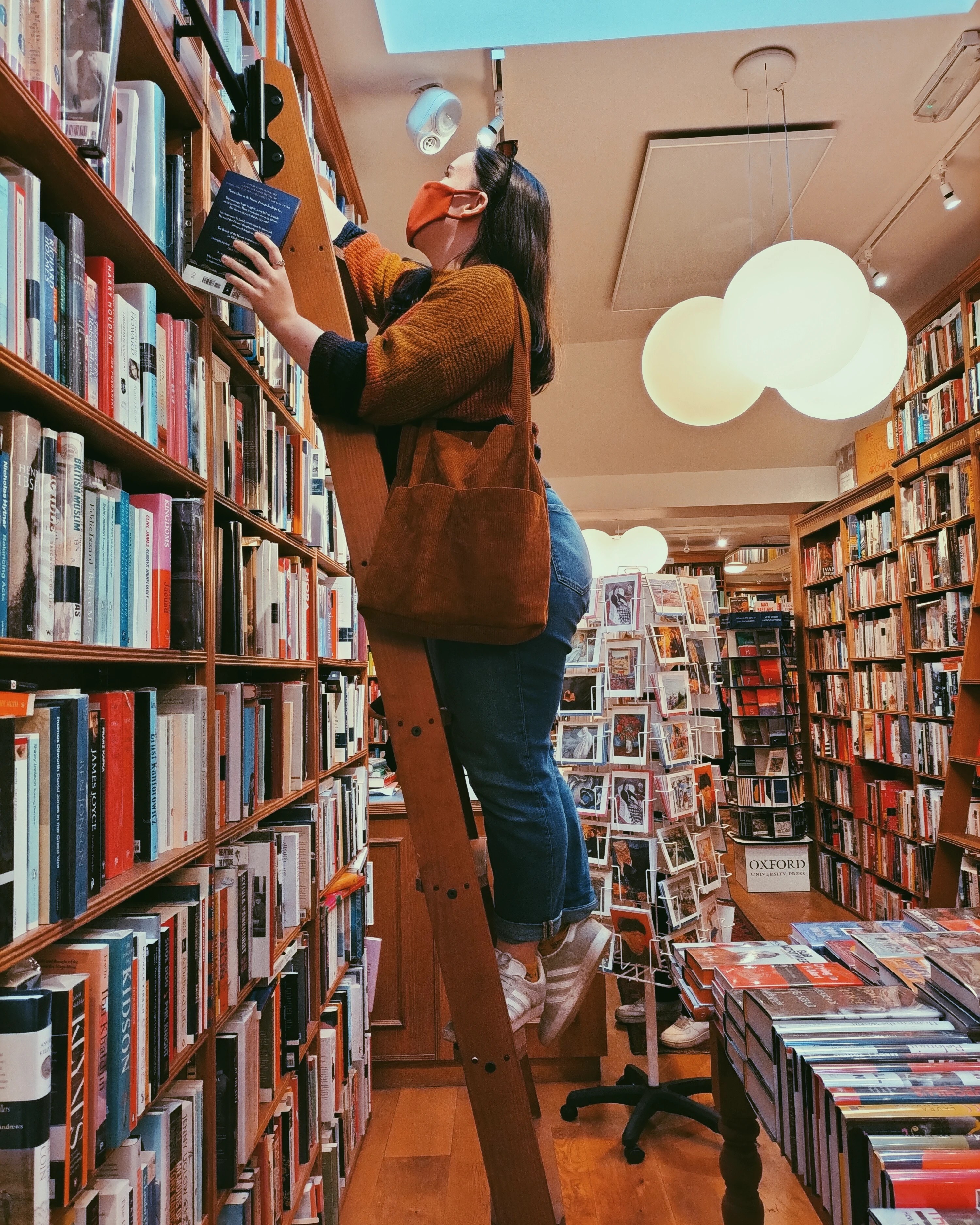 A small woman stands on a ladder looking at books on a bookshelf