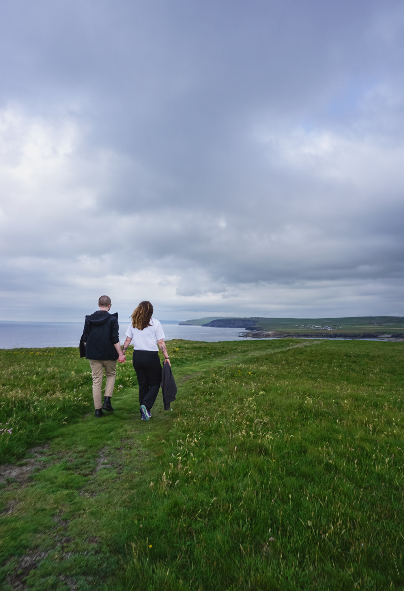 A couple walk together outside, they are on the grass, the sea is in the distance
