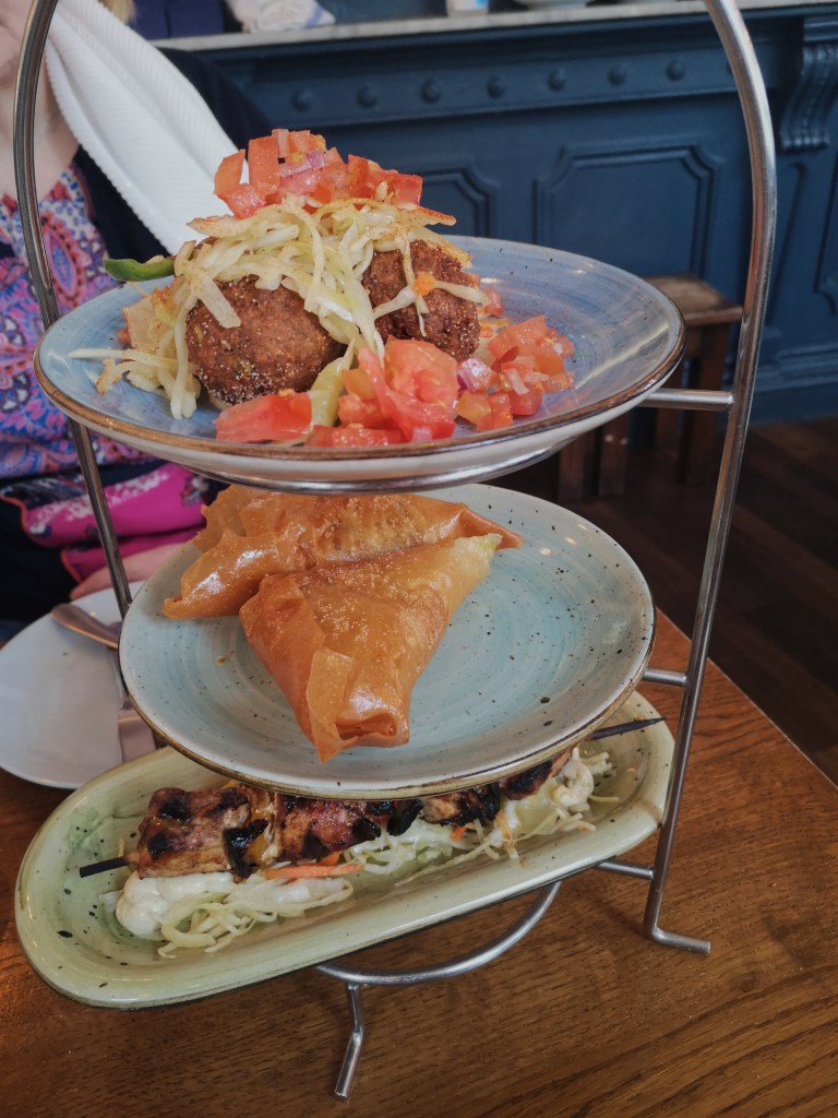 A stack of plates on a rack, one with a skewer of chicken, one with triangular filo parcels and the top has balls of falafel