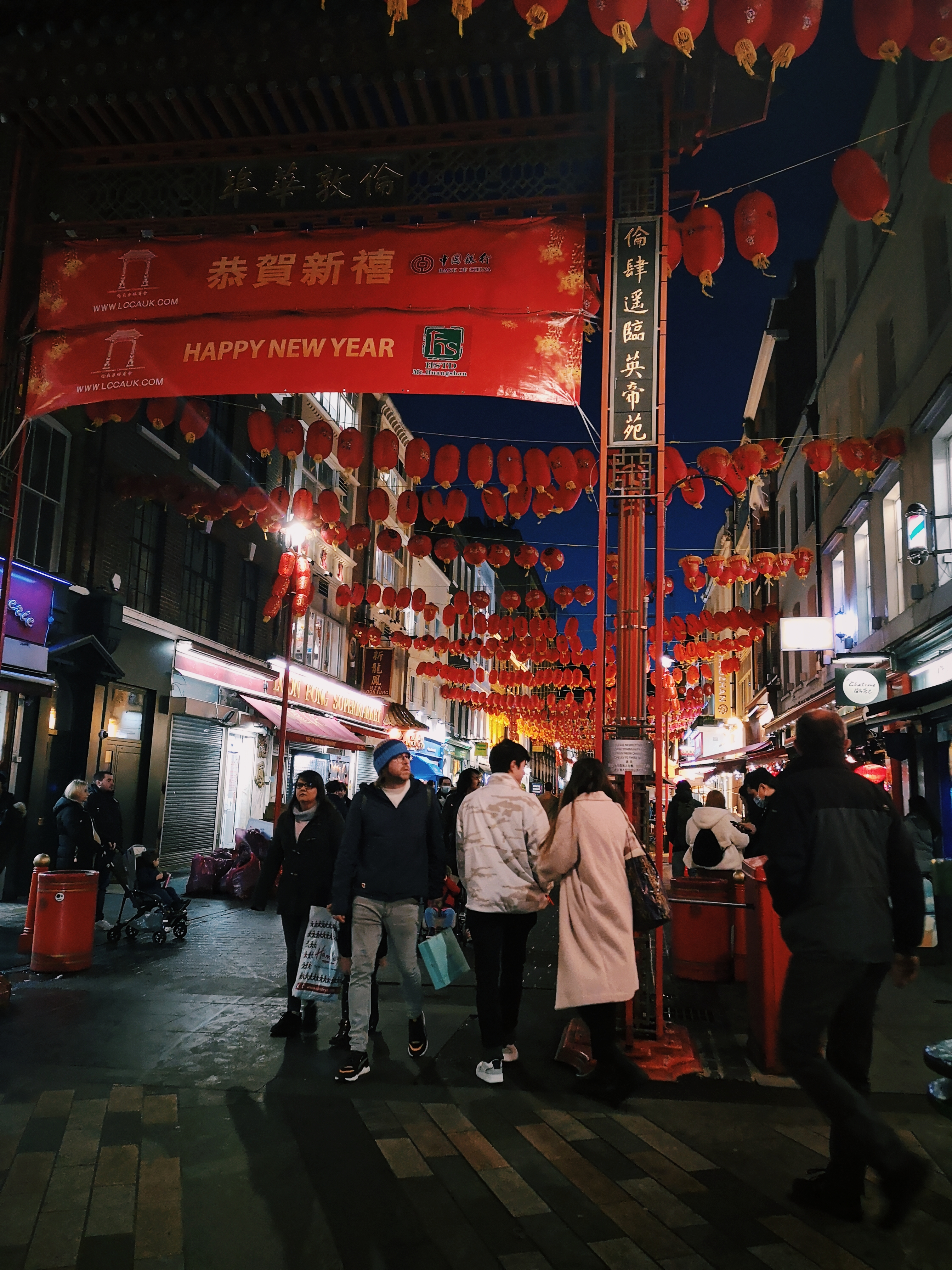 Signs and red lanterns are strung above a busy street. The sign says happy new year