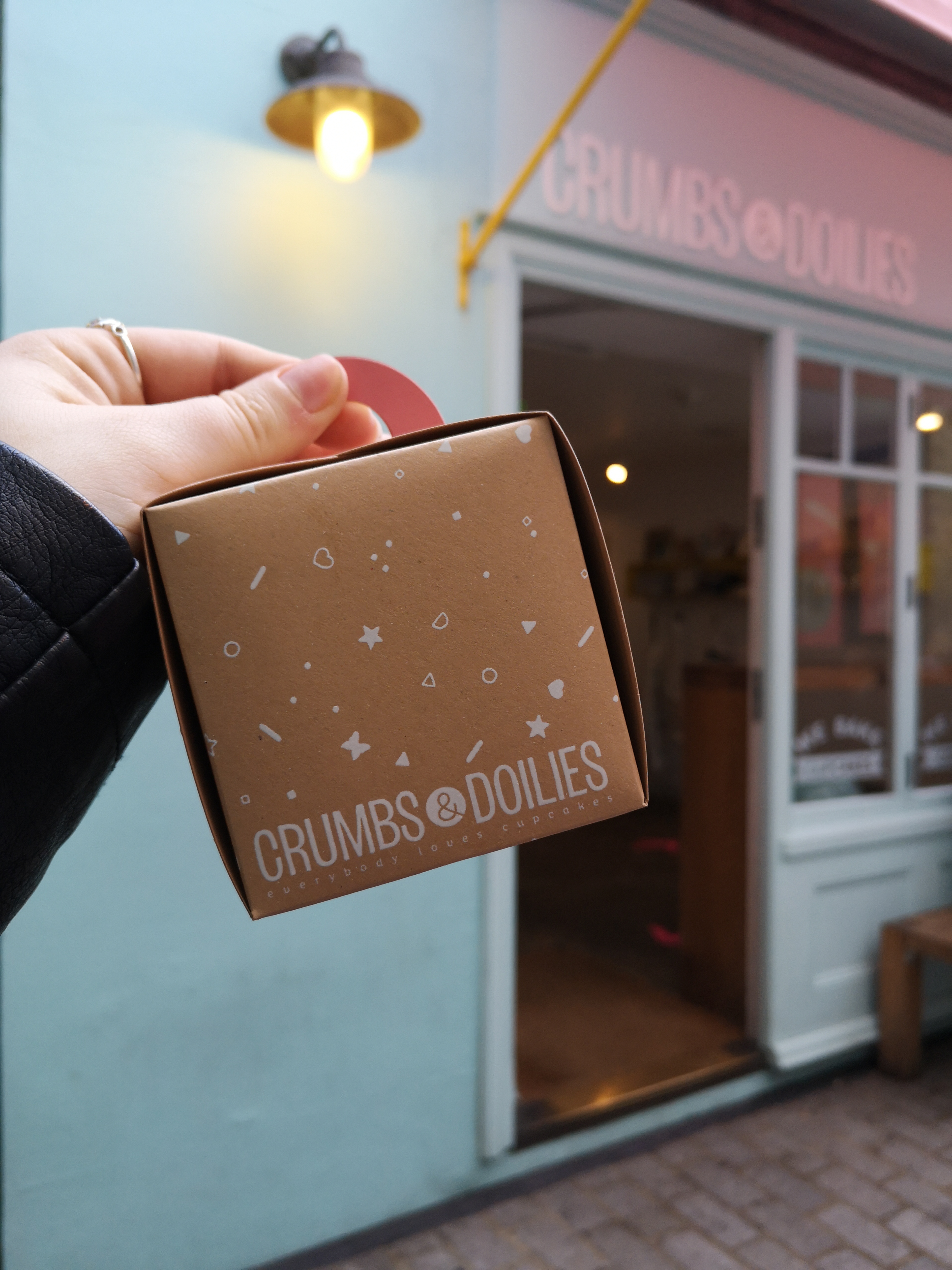 A little brown box that says Crumbs & Doilies is held up in front of a light blue shop front