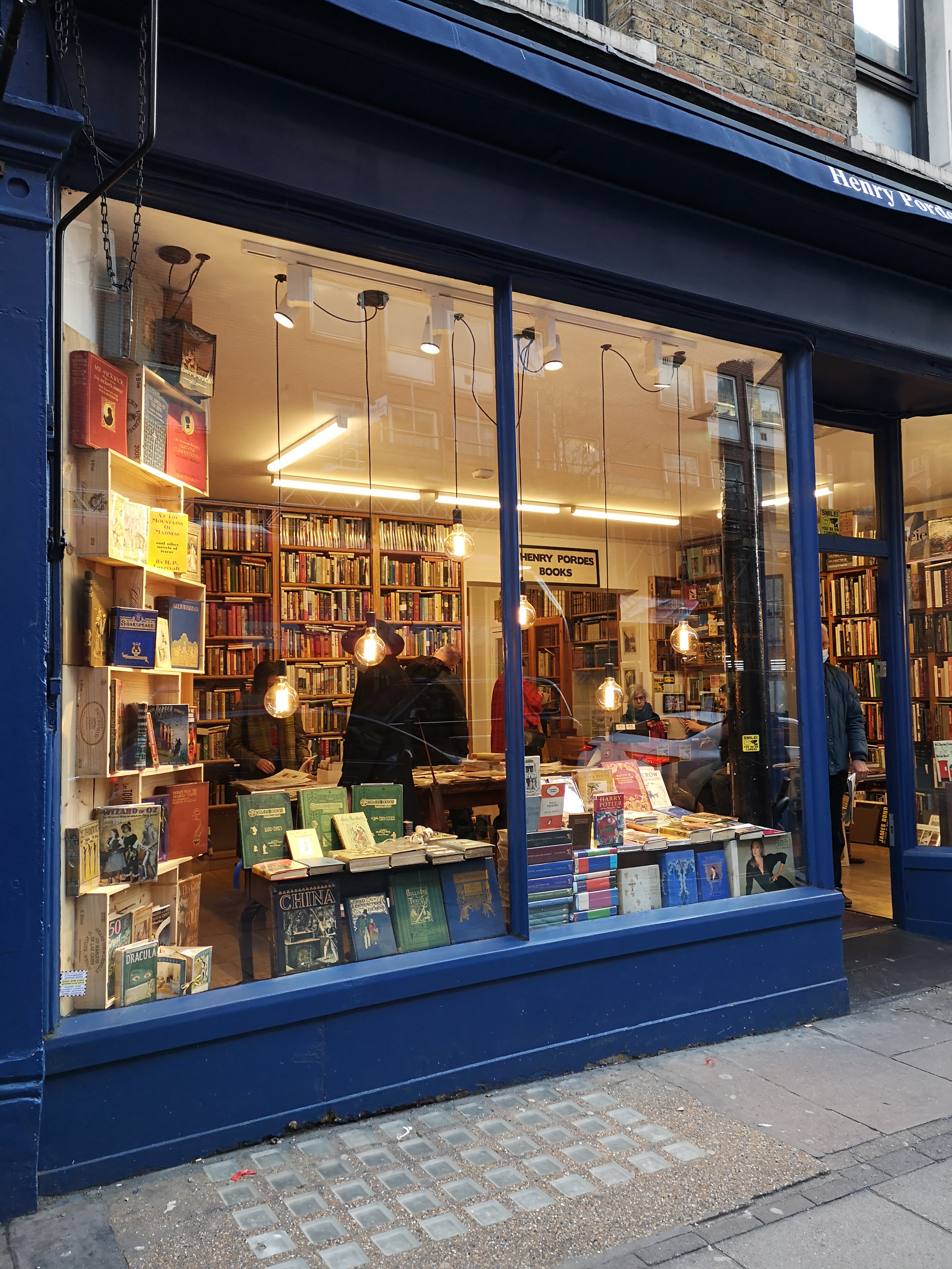 The navy shop front of a cosy bookshop, stacks and shelves of books behind hanging pendant lights