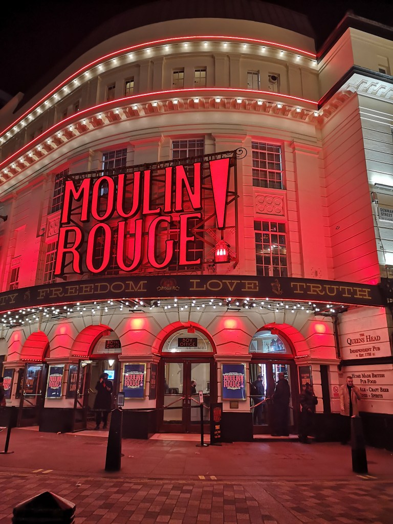 Outside of the Piccadilly Theatre with lights illuminating the buiding and a big red sign that says Moulin Rouge!