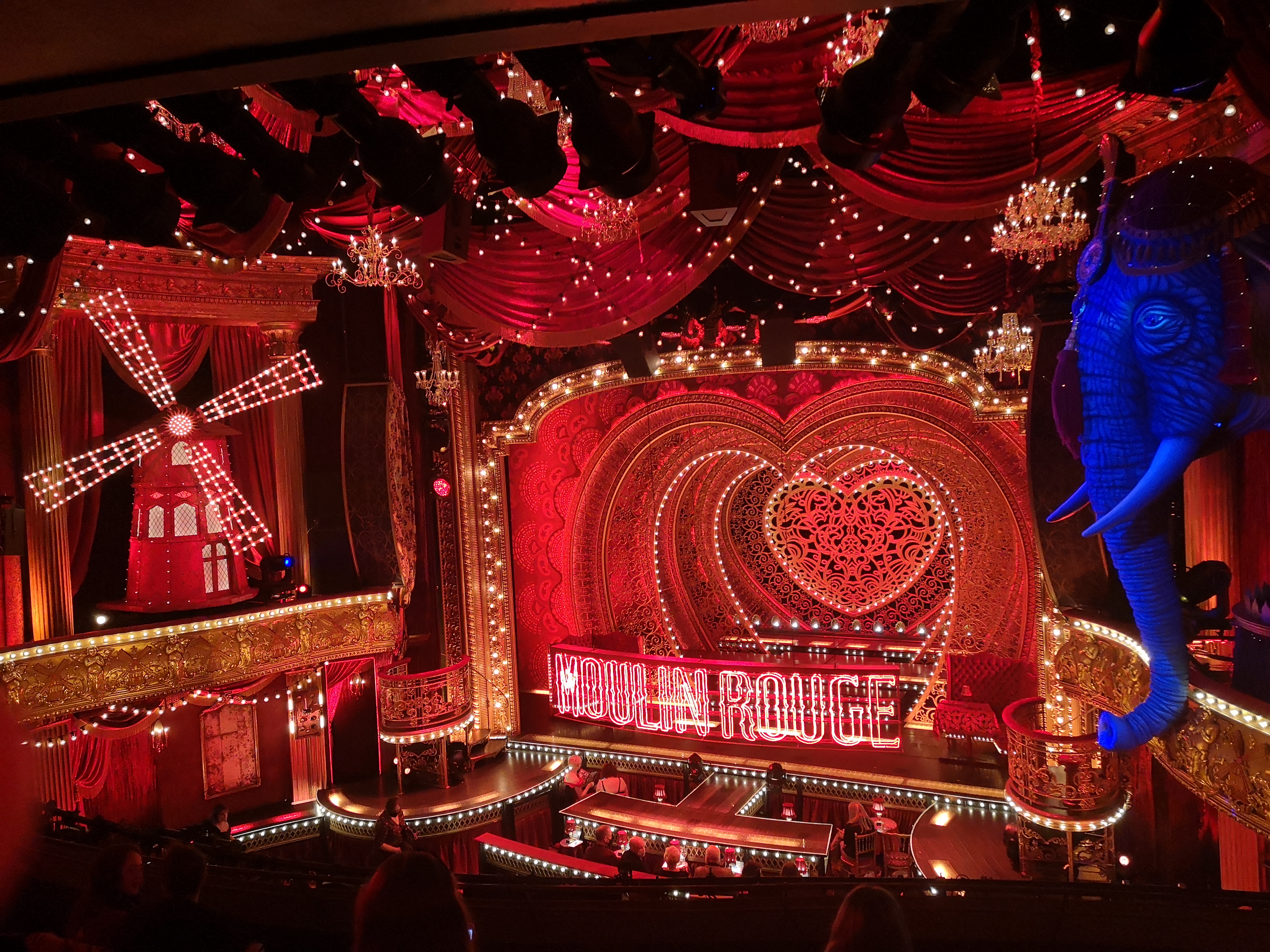 The ridiculously beautiful inside of the Piccadilly Theatre. There are white and red lights everywhere. The stage has layers of patterned stars and a big sign that says Moulin Rouge. To the left of the stage is a big red windmill and to the right is a blue illuminated elephant