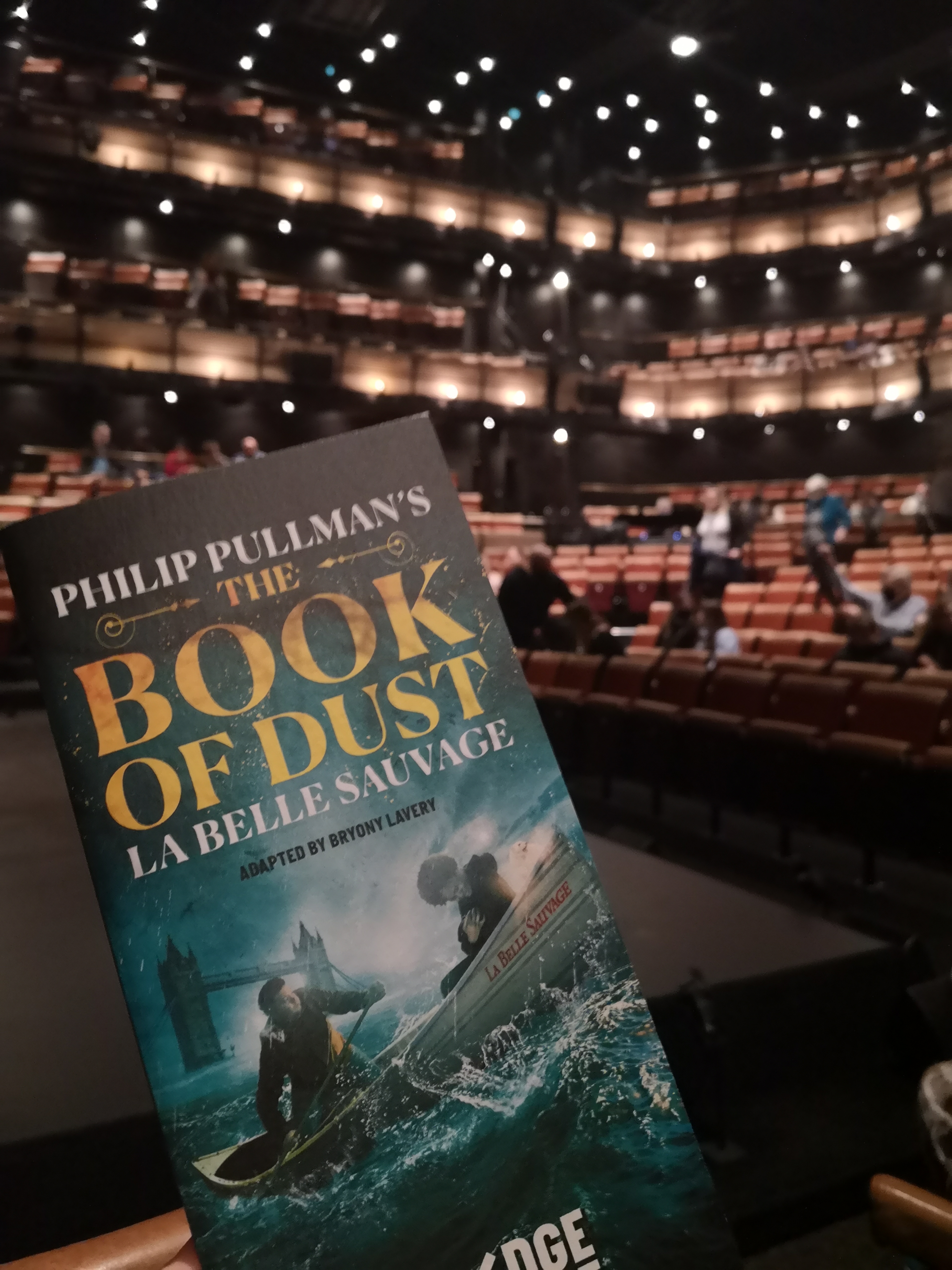 A programme with two people on a boat and the words Phillip Pullman's The Book of Dust La Belle Sauvage is held in front of a theatre, there are layers of balconies with strings of lights on them