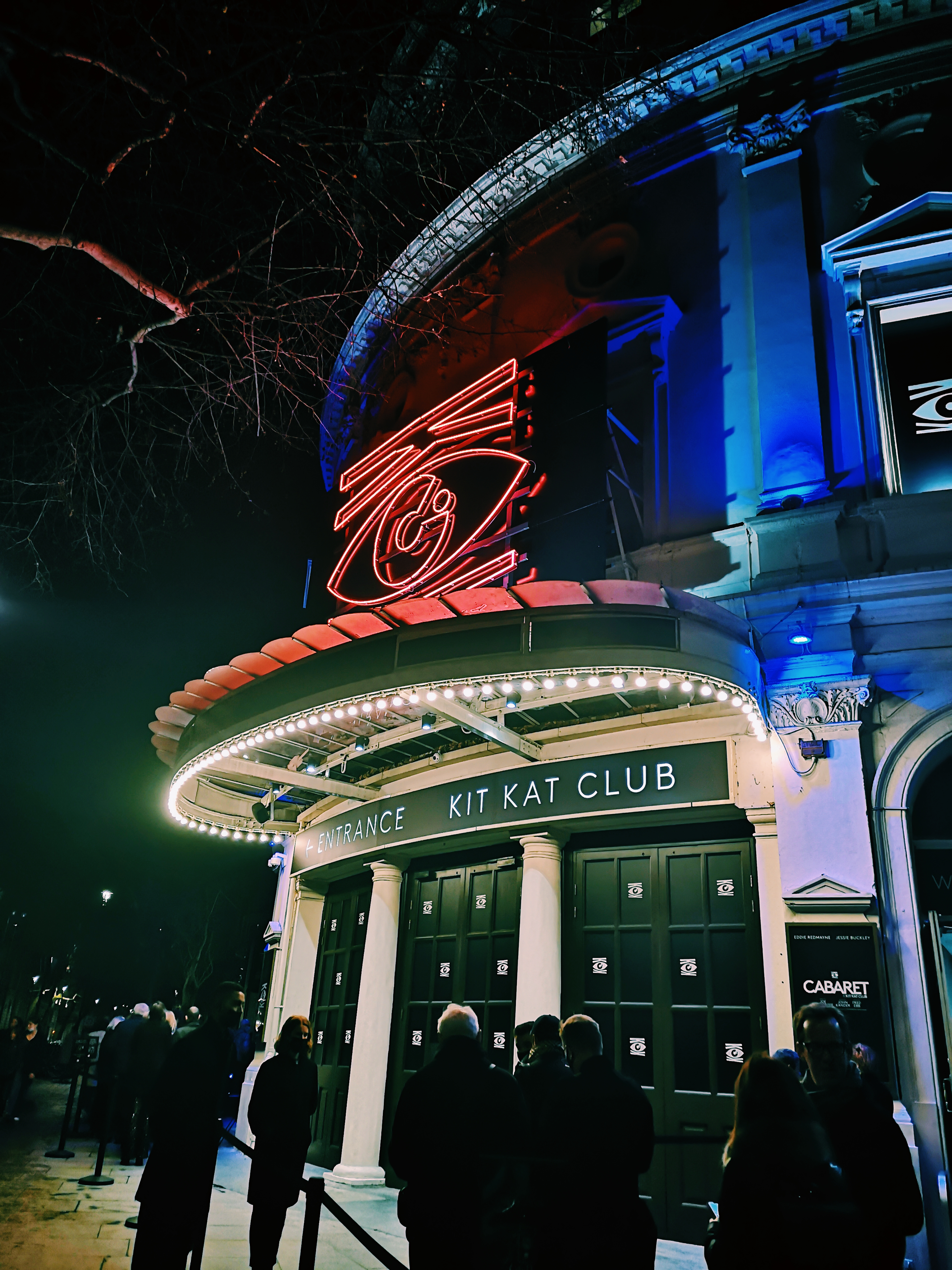Outside the Playhouse theatre, lights iluminate the sign that says Entrance to the Kit Kat Club and there is a red neon eye. 