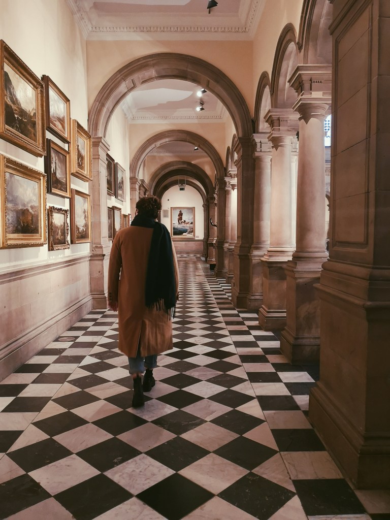 My friend Laura walks down an ornate corridor, arches curve in front of her, the floor is black and white check and there's paintings on the wall