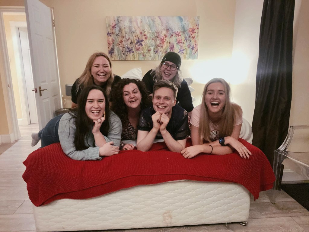 Six friends pile on top of each pother on a bed, laughing
