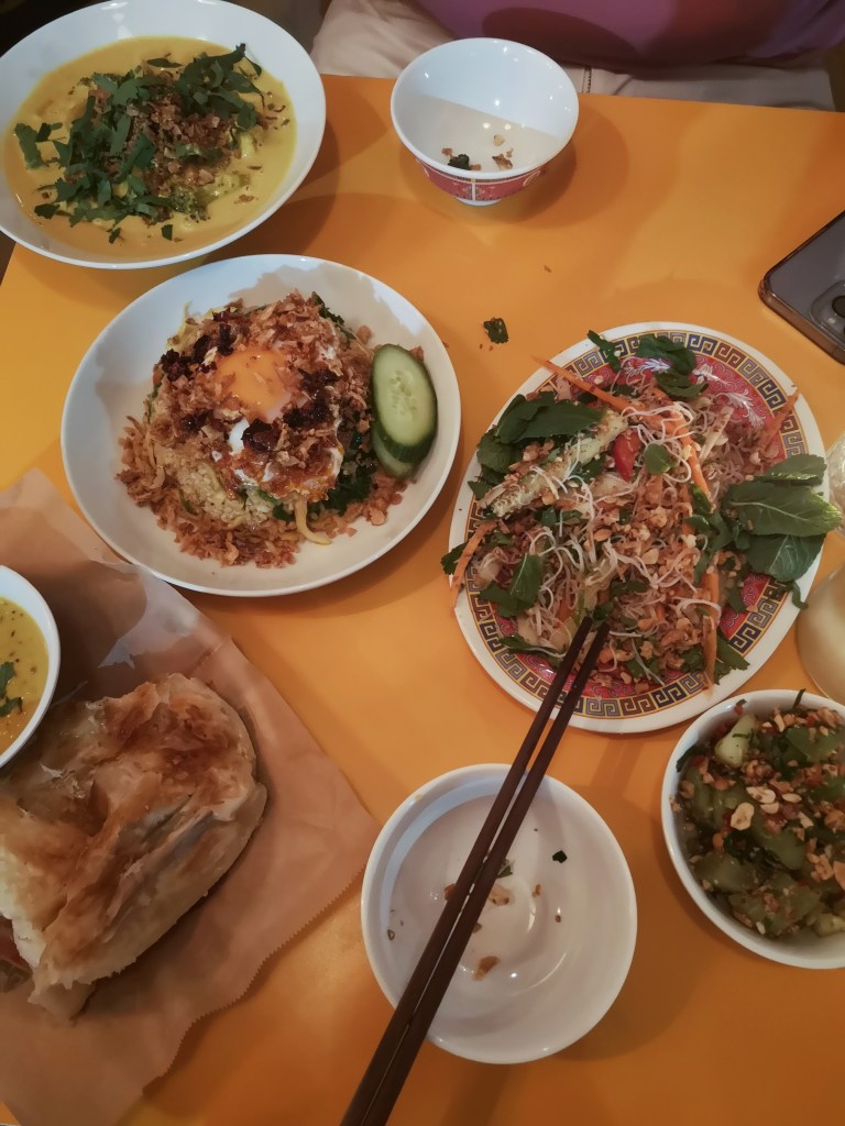 A table with many delicious dishes, a noodle salad, rice with a fried egg on top, roti breads, a yellow curry