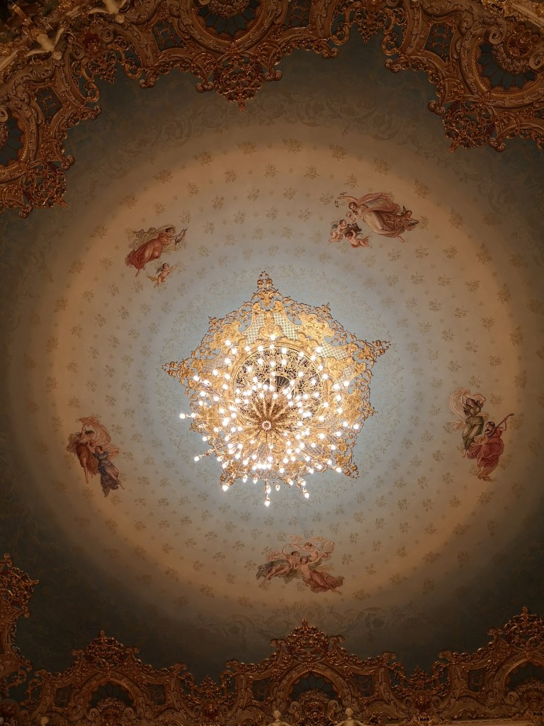 The ceiling of the Teatro Al Fenice with a gold ornate chandelier and cherubs painted 