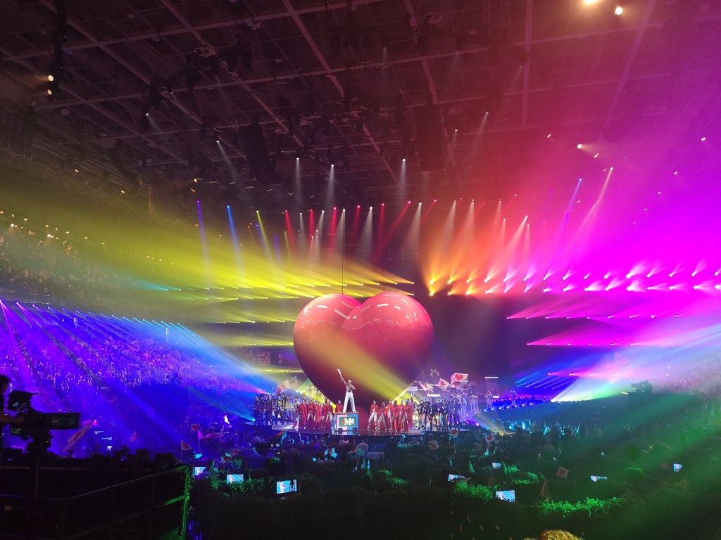 Colourful lights and a massive red heart on stage behind legend and icon Mika who stands on a box with an arm raised