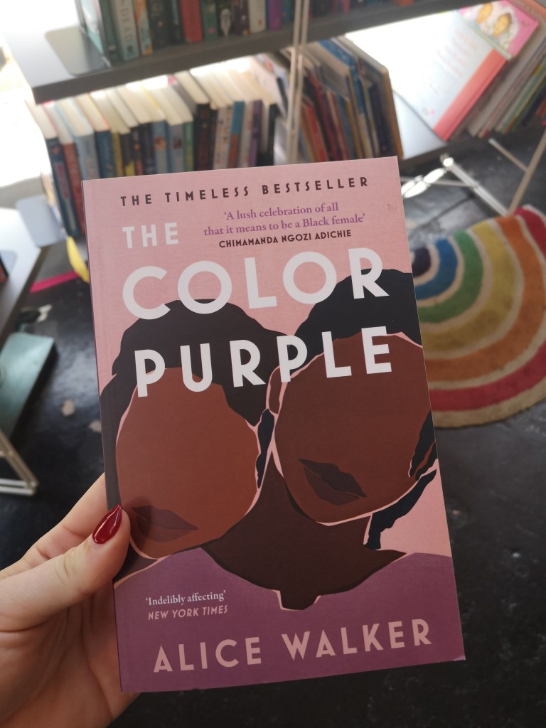 A pale pink book held in front of a small rug shaped like a rainbow. There is a graphic illustration of two black women leaning on one another