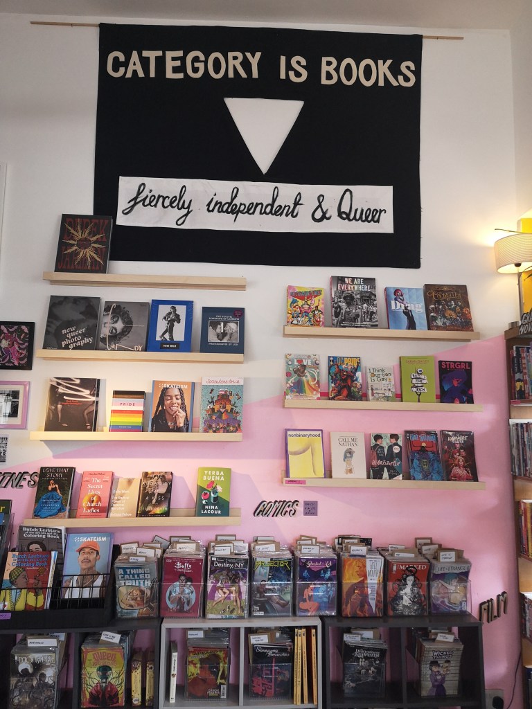 The interior of Category Is Books, there are flaoting shelves highlighting favourite books and comics. There is a big sign on the wall that says 'fiercely independent & queer'