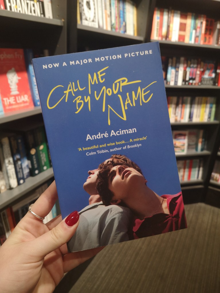 A blue book with yellow text that reads Call Me By Your Name held up in front of a bookshelf. On the cover two men lean their heads against one another tenderly.