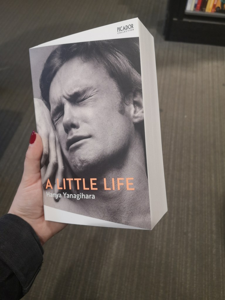 A man closes his eyes wincing with his hand held beside his face on a very chunky book called A Little Life