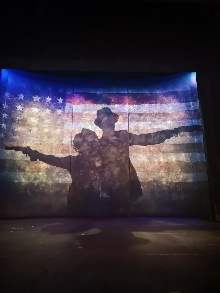 A silhouette of Bonnie and Clyde holding guns out to either side is projected on top of an American flag