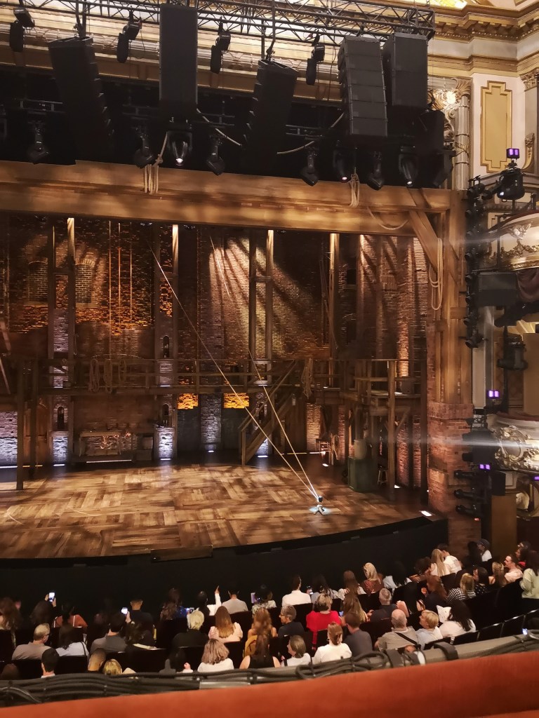The stage of Hamilton is a lot of wood, there is a platform with a handrail and two ropes cross from the ceiling to hook around a spot on the floor
