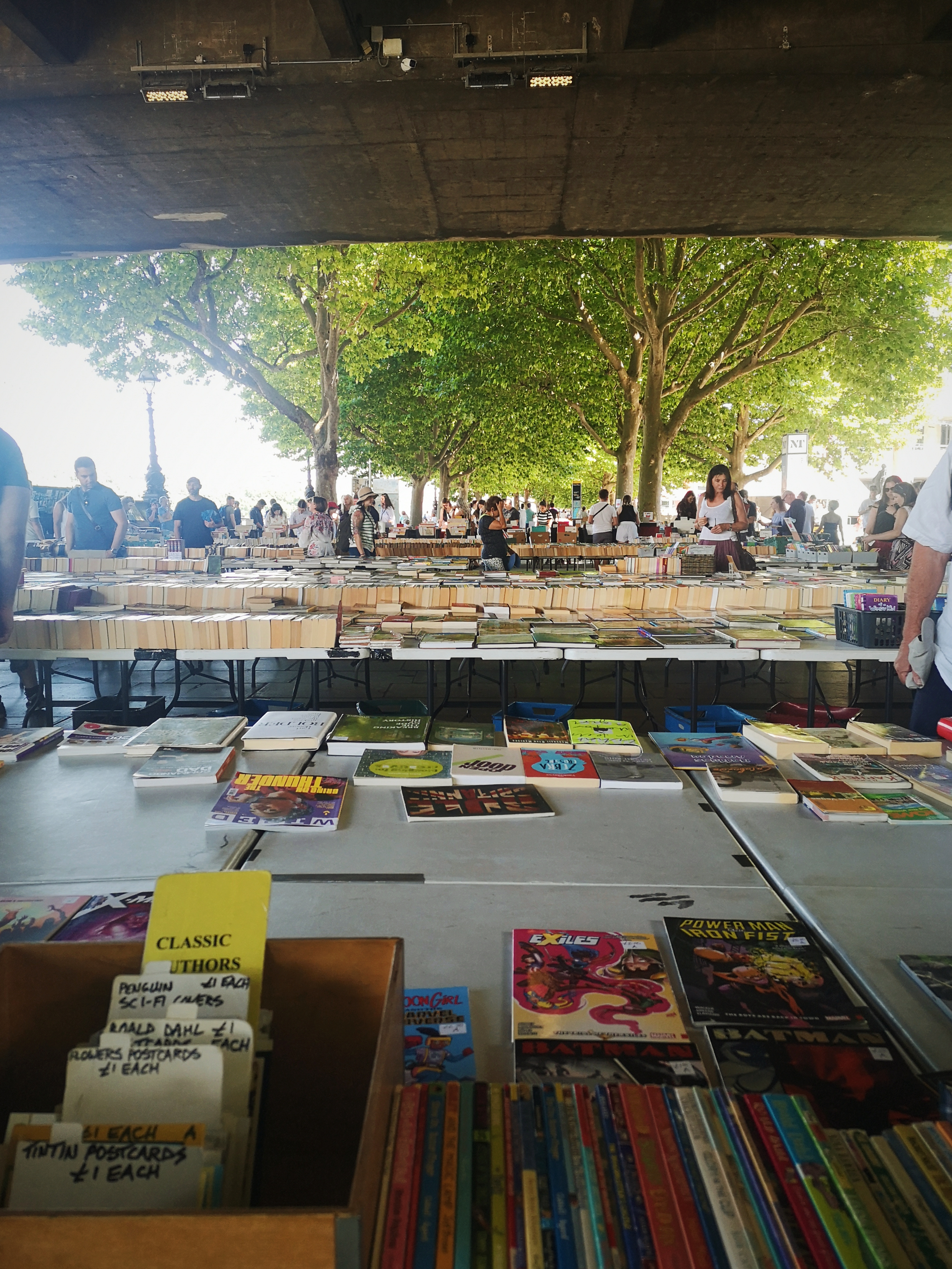 Multiple tables full of books underneath a bridge in London with trees in the background