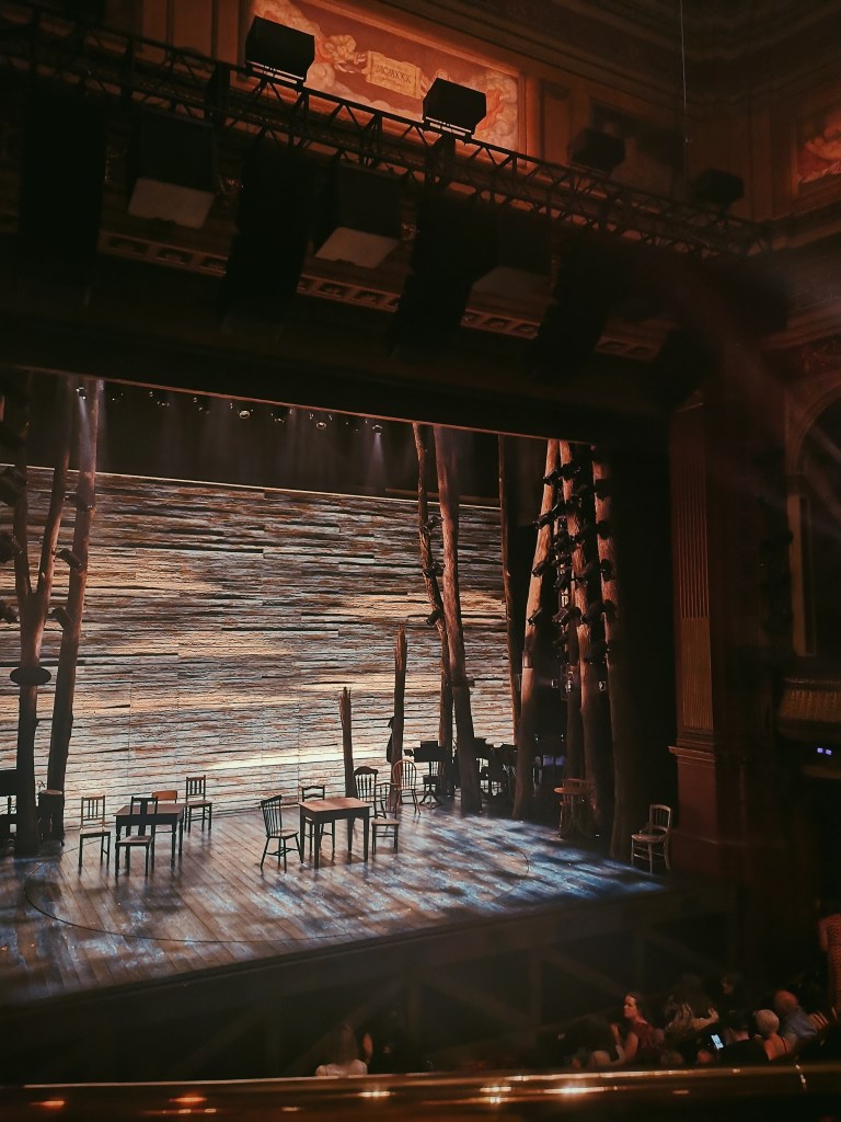 The stage of Come From Away has tables and chairs and the trunks of trees lined up at the edges, again there is a lot of wood
