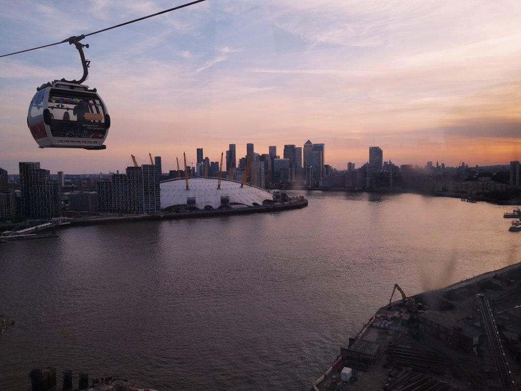 A cable car hangs over the river Thames with the O2 arena and a skyline in the background, the sky is orange with the sunset