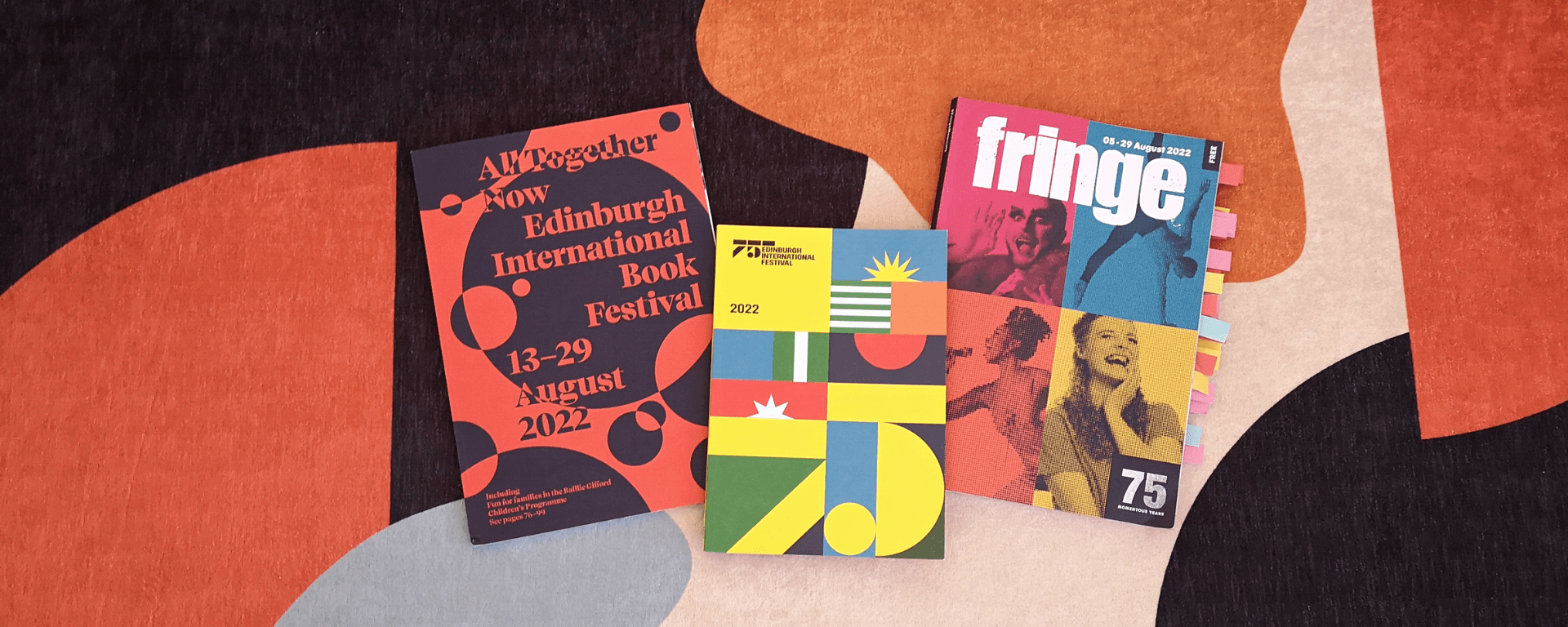 Three brochures sit on top of a colourful rug, they are advertising the Edinburgh Book, International and Fringe festivals