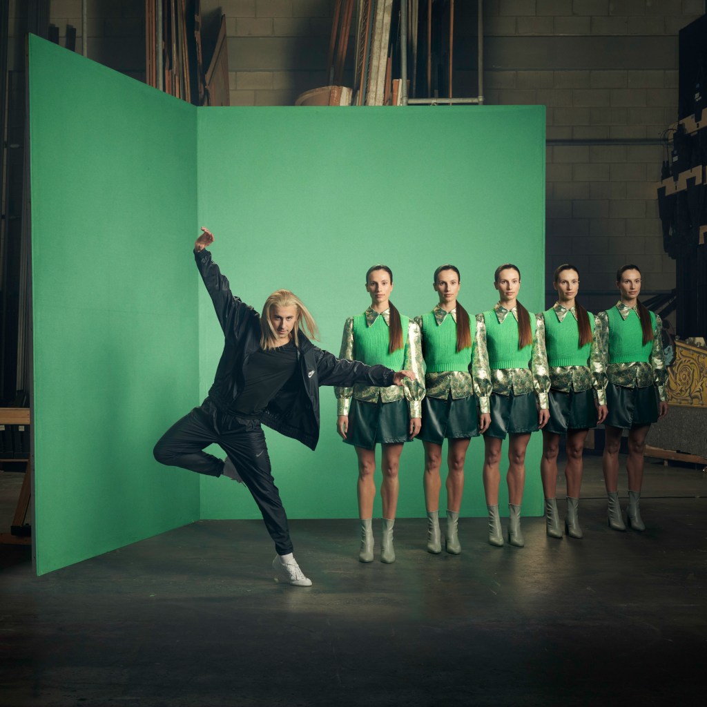 A man in a black tracksuit dances, one leg bent with his foot behind the other knee and his arms stretched out in a sort of L shape. Behind him is 5 cloned women standing like robots with a straight face. They all stand in front of a big green wall.